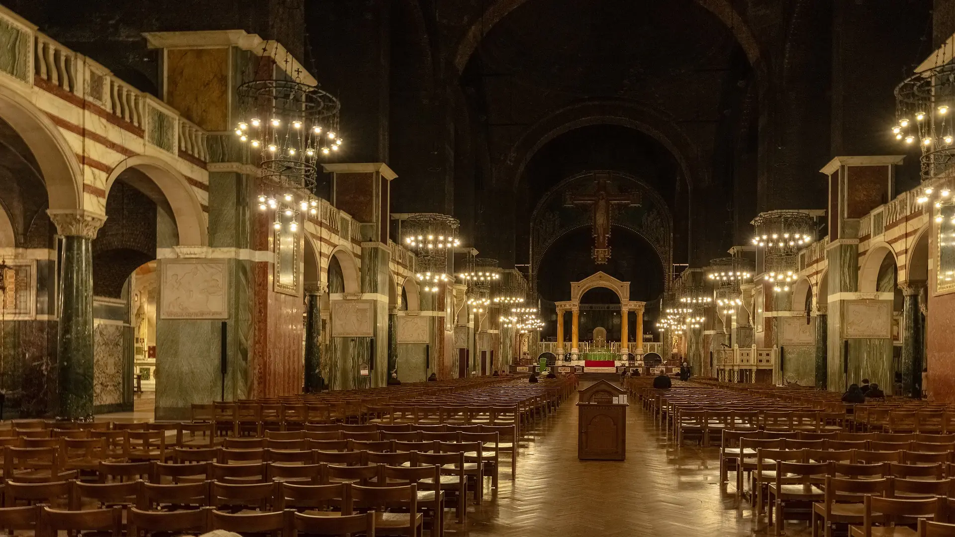La nave de Westminster Cathedral de noche, con vistas hacia el este más allá de las arcadas de mármol rayado y las lámparas colgantes, hasta la gran cruz de Rood y el baldaquino sobre el altar mayor.